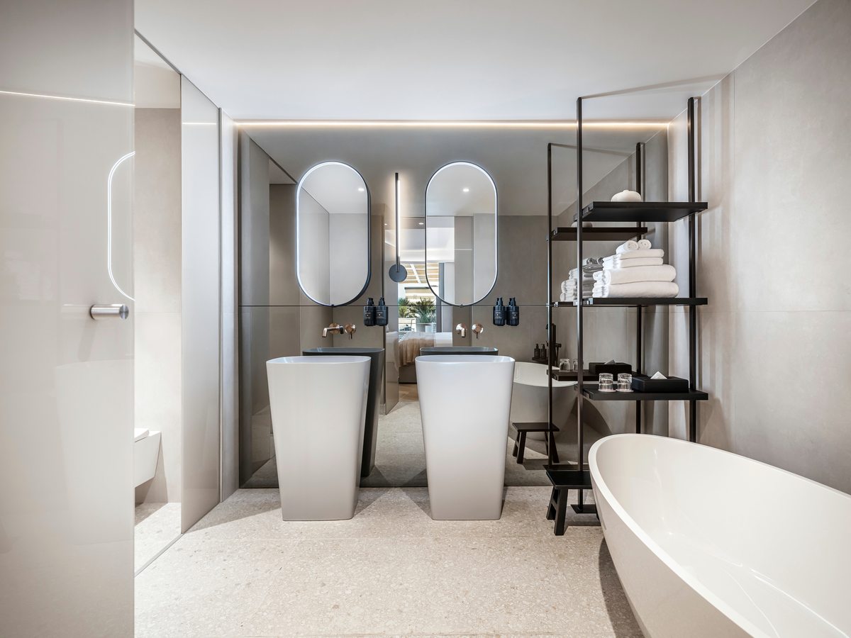 Room bathroom with twin freestanding basins under backlit oval mirrors, open towel shelving, a walk-in shower and a bath.