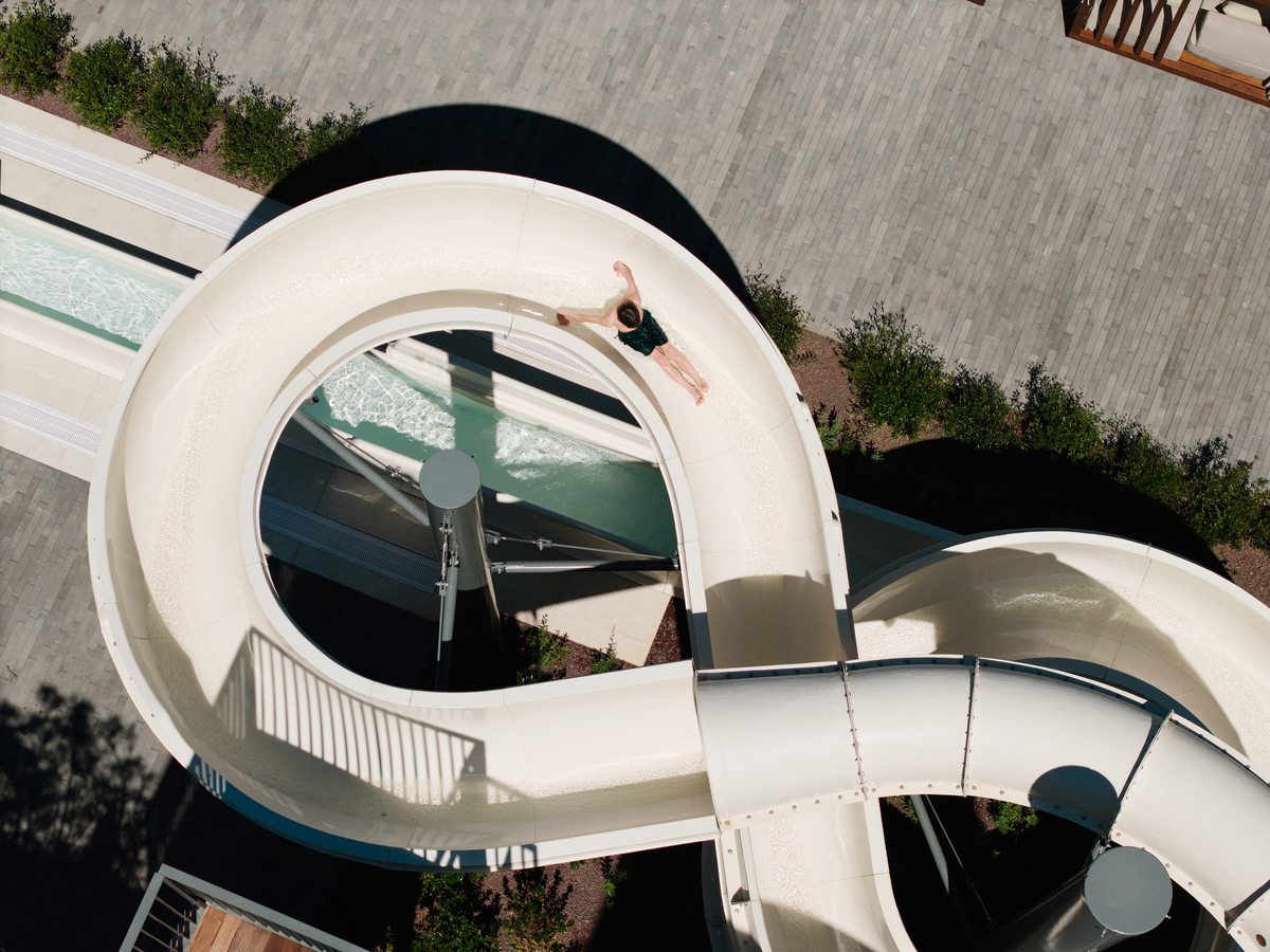 Two white water flumes looping above a paved poolside with planted borders, and an adult sliding down one of them.
