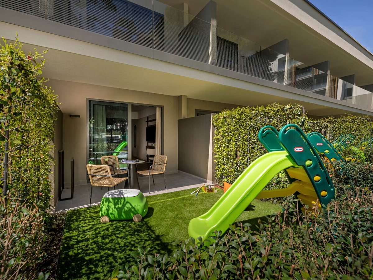 Ground-floor room's garden terrace with a table and rattan chairs on paving, artificial grass, a toddler slide and a ride-on toy, screened by a tall hedge.