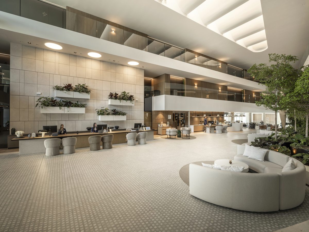 Multi-storey atrium lobby with reception desks along one wall, a large circular white sofa, tall indoor plants and balcony corridors above.
