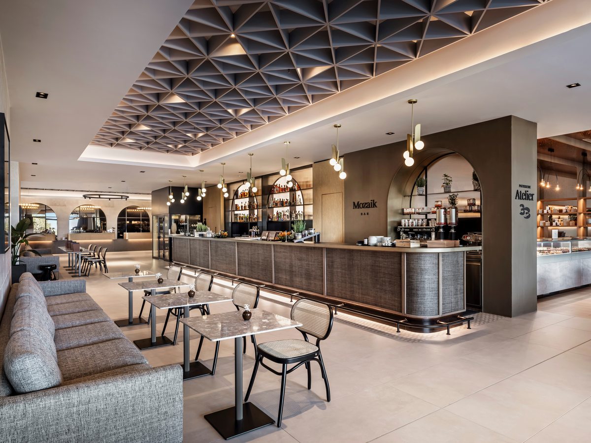 Long lobby bar counter with bottle shelving and brass pendant lights, small marble tables and cane chairs in front, and a patisserie counter through an arch to the right.