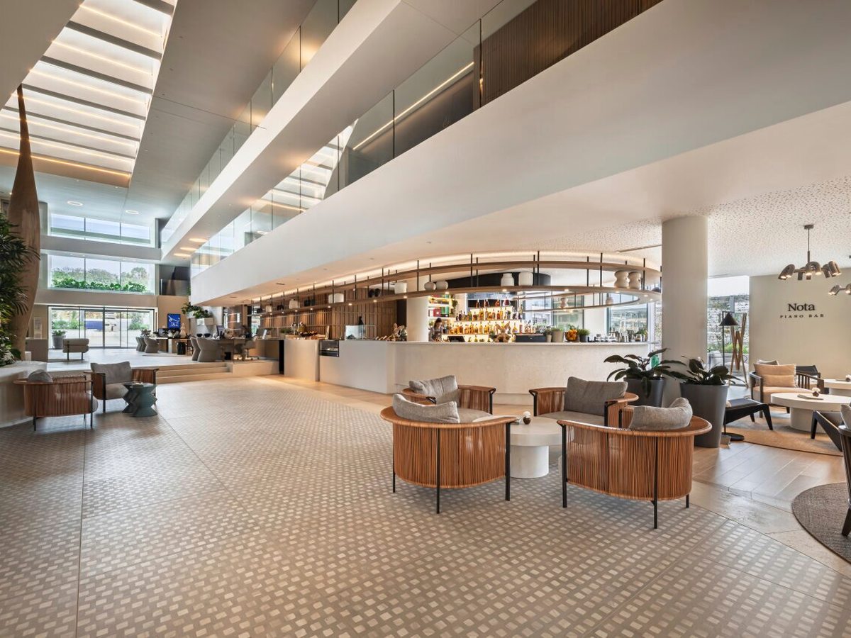 Curved bar counter in a double-height lobby with a glazed roof, rattan tub chairs on a patterned tile floor and a gallery landing above.