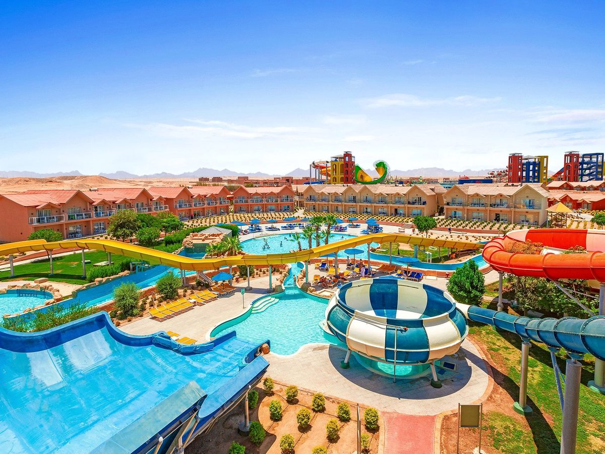 A wide view of the aqua park, with a lazy river curving between palms, coloured slides, a domed building and terracotta hotel blocks beyond.