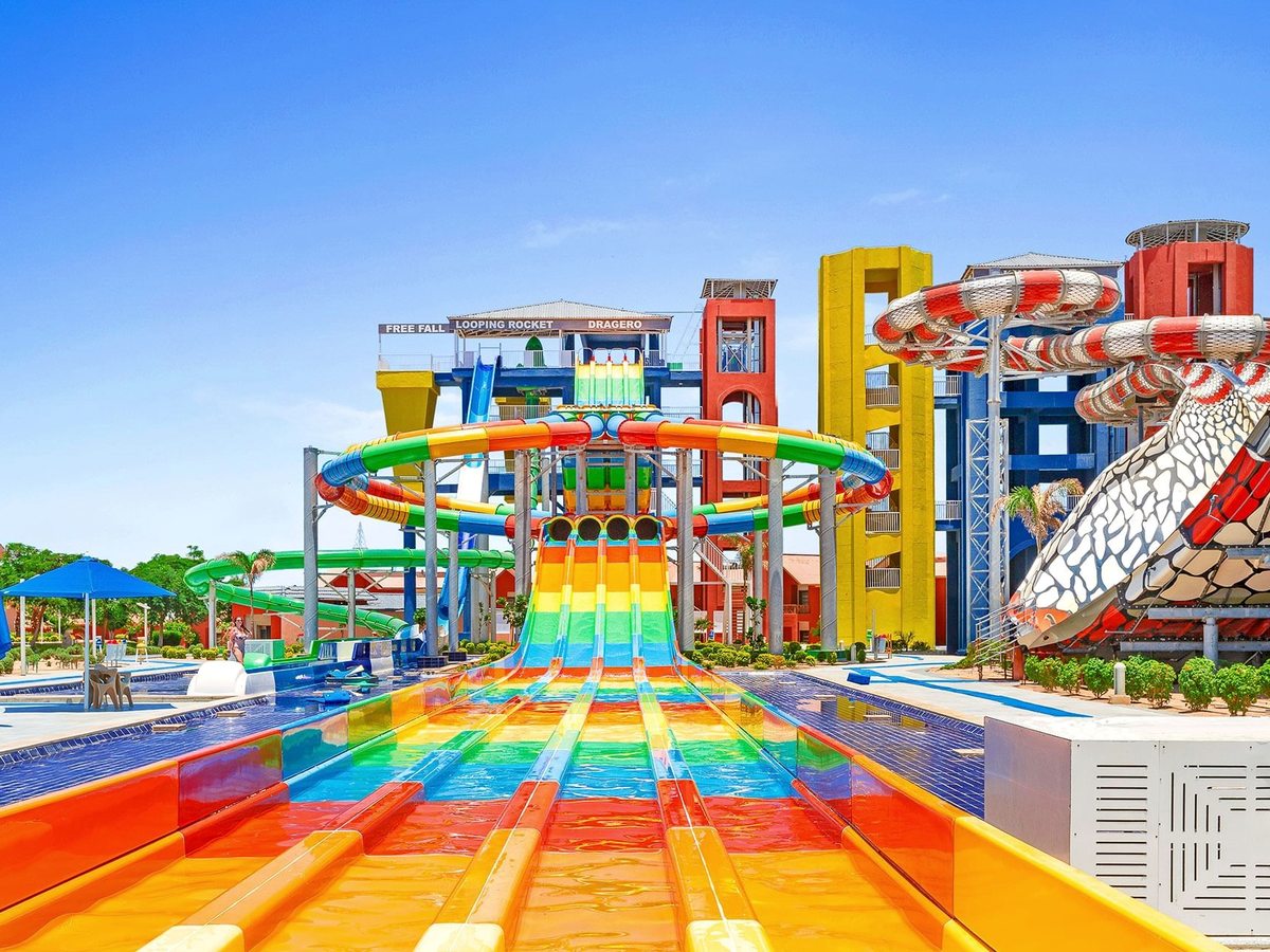 Rainbow-coloured racing slides side by side running down to a landing lane, with a tall slide tower and looping tube slides behind.