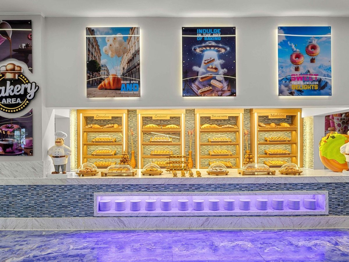 A bakery counter with lit wooden shelves of croissants, cinnamon buns and madeleines behind a marble top, under three backlit posters.