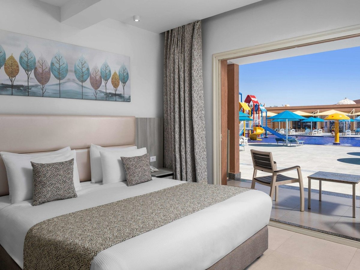 A grey and cream double bedroom with a leaf painting above the bed, opening onto a terrace beside a pool with children's play structures.