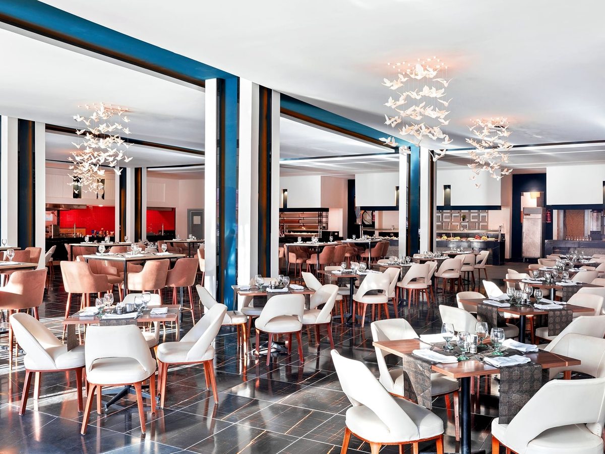 A restaurant with blue and white columns, white and pink chairs at laid tables, bird-shaped chandeliers and an open kitchen counter along the back.