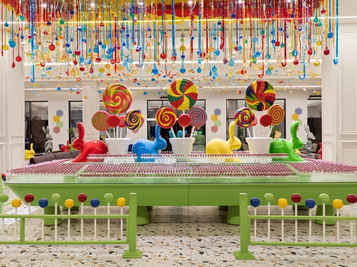 A green lobby counter covered in rows of pink lollipops, with giant swirl lollipop models, coloured rabbit sculptures and thousands of hanging glass sweets above.