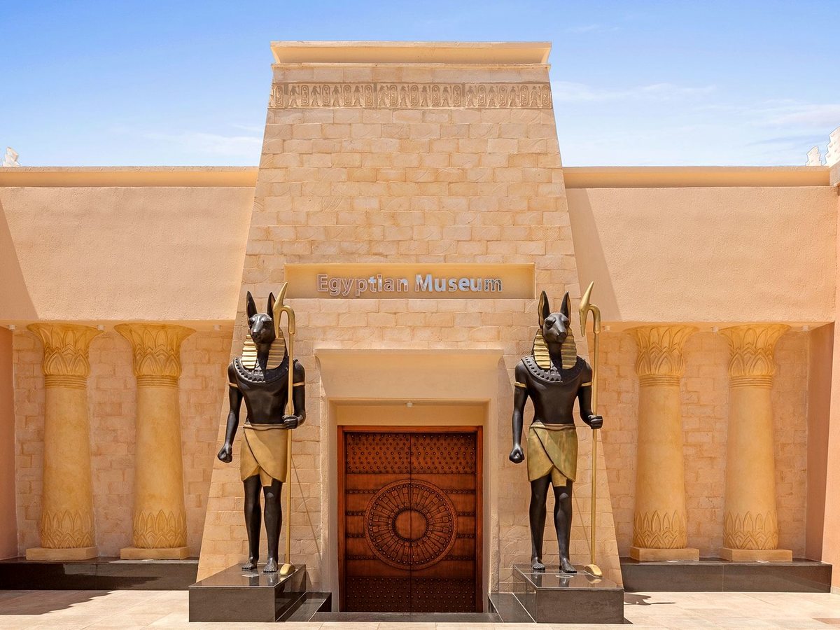 A sandstone temple front with carved lotus columns, an Egyptian Museum sign and two black jackal-headed statues either side of a wooden door.