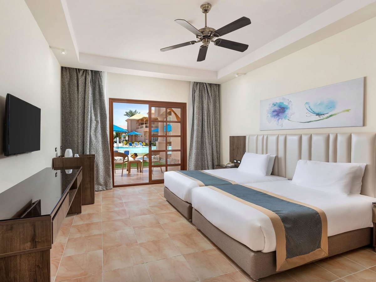A twin bedroom with tiled floor, ceiling fan and a wall-mounted television, with wooden French doors open onto a terrace and a pool beyond.
