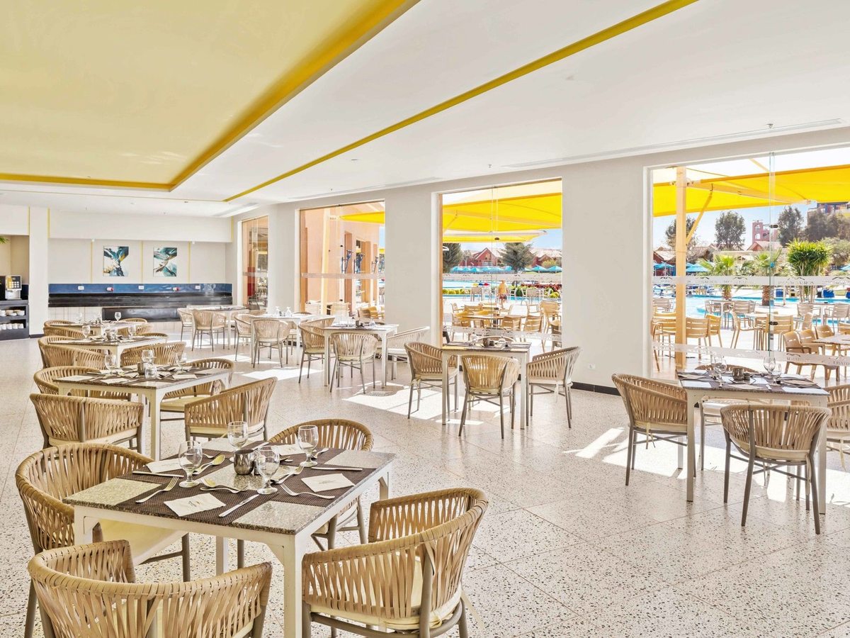 A bright dining room with cream woven chairs and laid tables, opening through full-height windows onto a terrace of yellow parasols beside a pool.