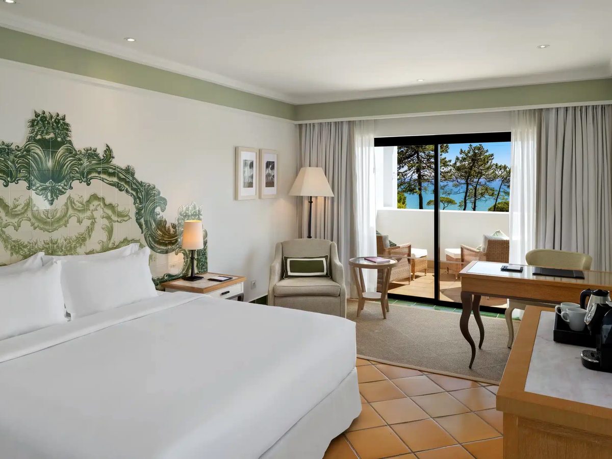 A double room with a painted green and white ceramic headboard, terracotta floor, armchair and writing desk, and a balcony looking out to pines and the sea.