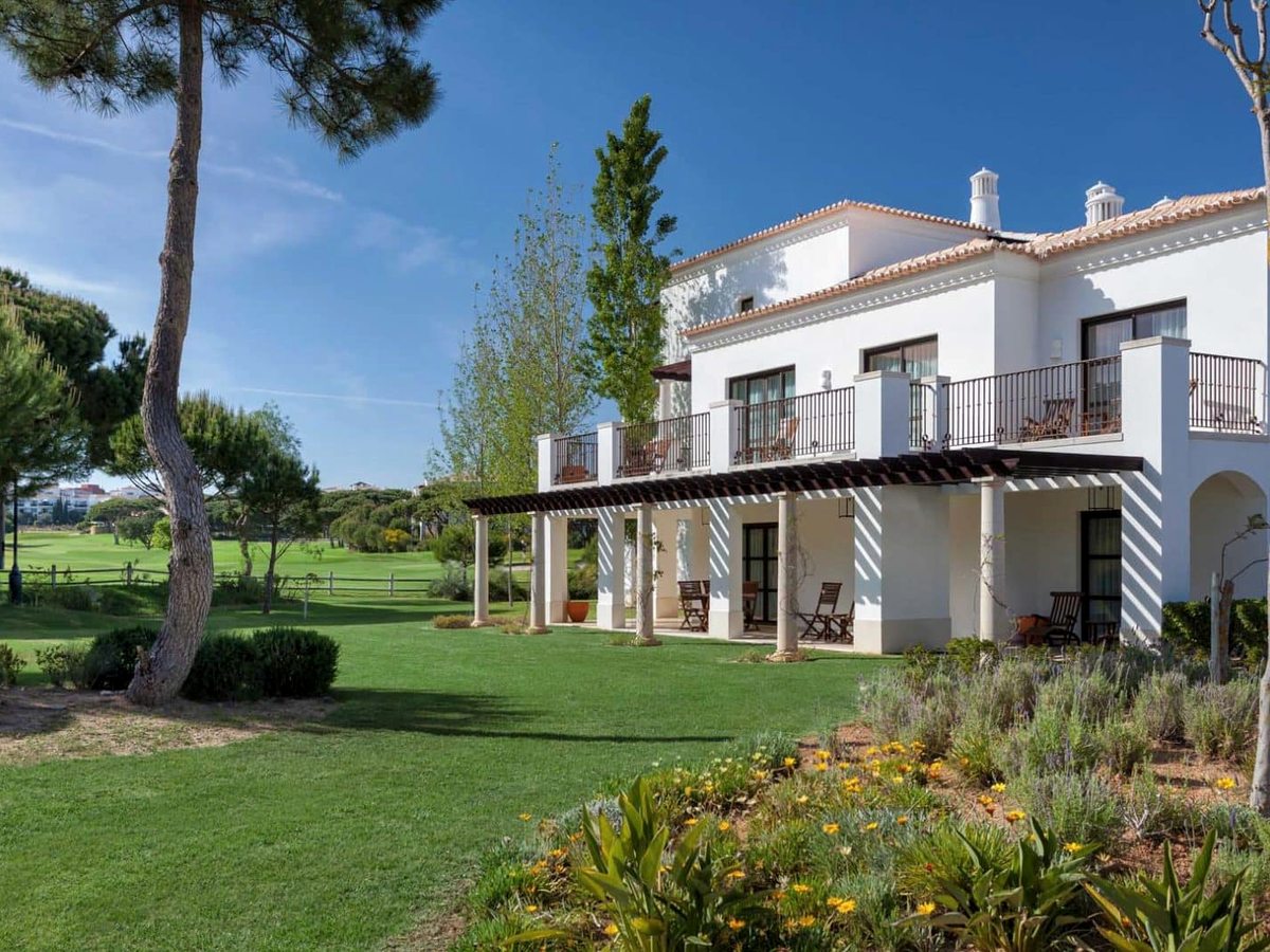 A white two-storey building with a tiled roof, balconies and a pillared veranda, on a lawn with flower beds, with the golf course and pines behind.