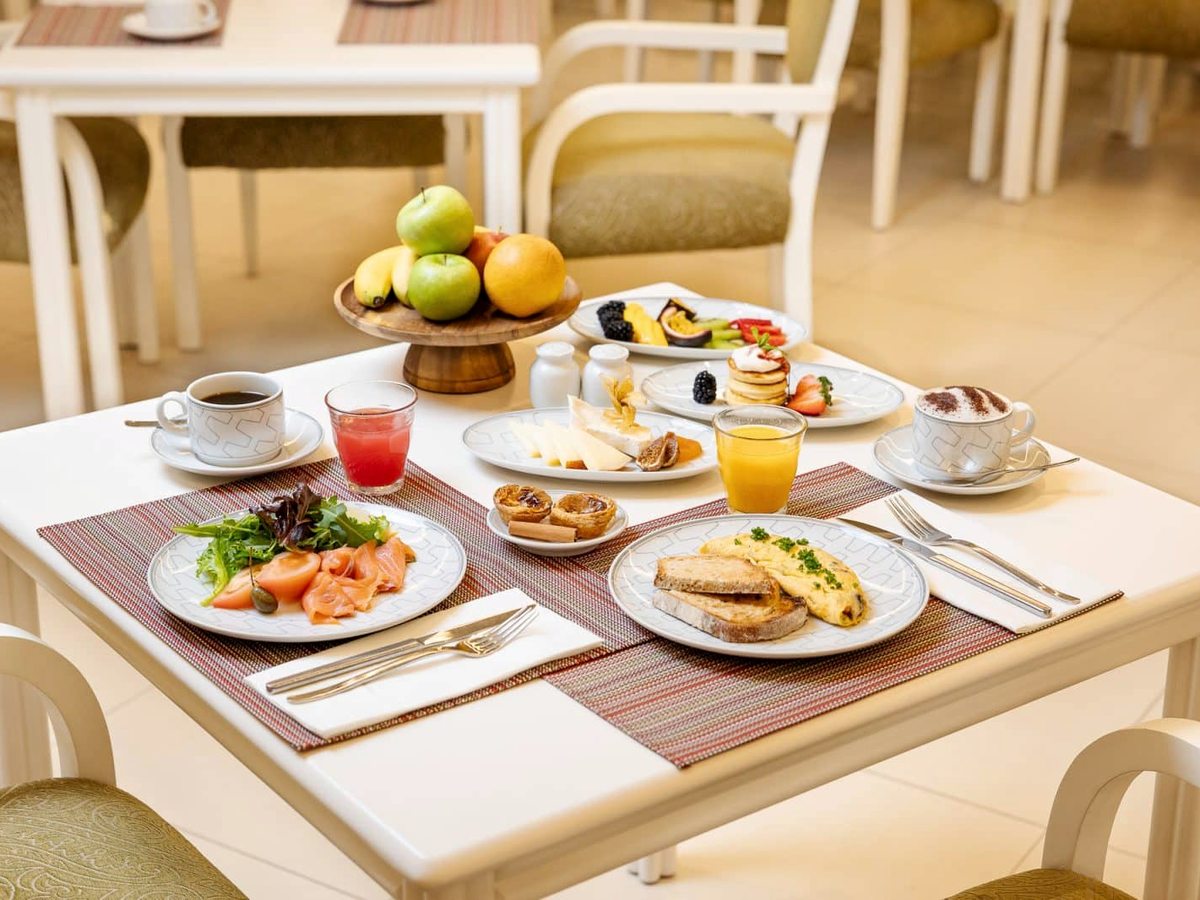 A breakfast table laid with coffee, orange juice, bread, pastries, fruit, eggs and smoked salmon in a light dining room.