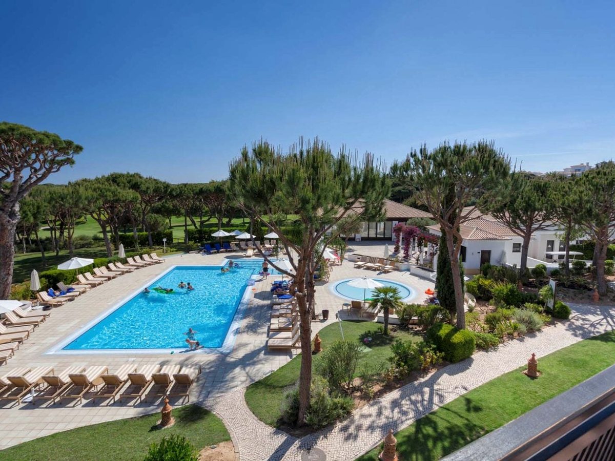 A rectangular pool with a round shallow pool beside it, rows of sun loungers, umbrella pines and the golf course behind.