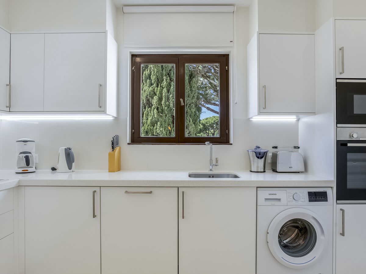A white fitted kitchen with a sink under the window, oven, microwave, kettle, toaster and a washing machine under the counter.