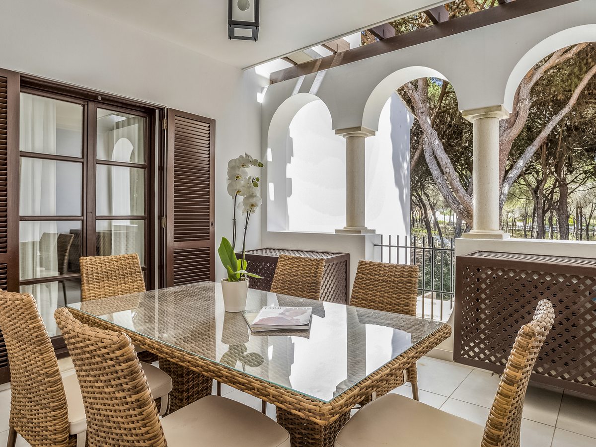 A covered terrace with a glass-topped dining table and six wicker chairs, arched openings and umbrella pines beyond.