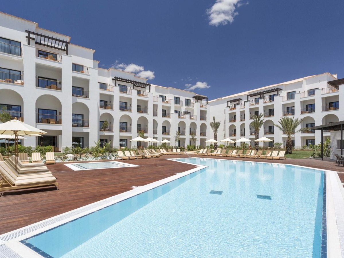 A large rectangular pool on a wooden deck, with sun loungers and parasols in front of a white apartment building with arched balconies.