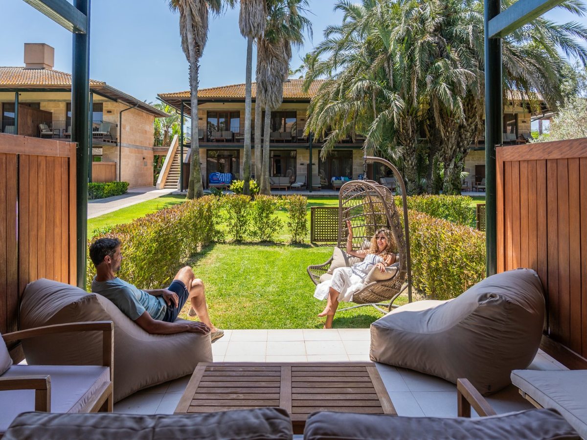 The view from a shaded room terrace with bean-bag seating out onto a private hedged lawn with a hanging egg chair, and two-storey resort buildings among palms beyond.