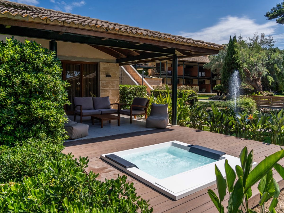 A small square pool set into a timber deck outside a tiled-roof room, with a covered lounge of sofas alongside and a garden pond and fountain beyond.