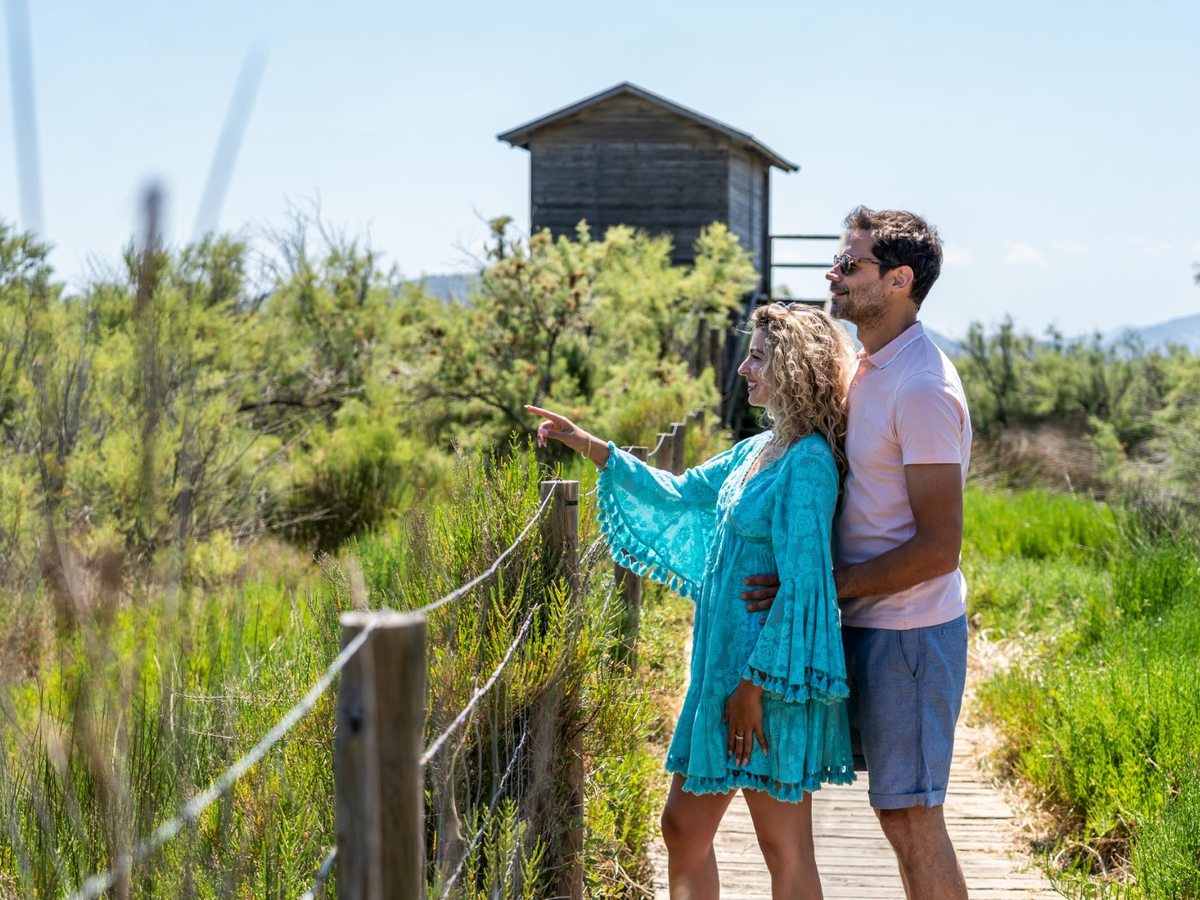A couple on a wooden boardwalk through low wetland scrub, with a raised timber bird hide behind them and hills in the distance.