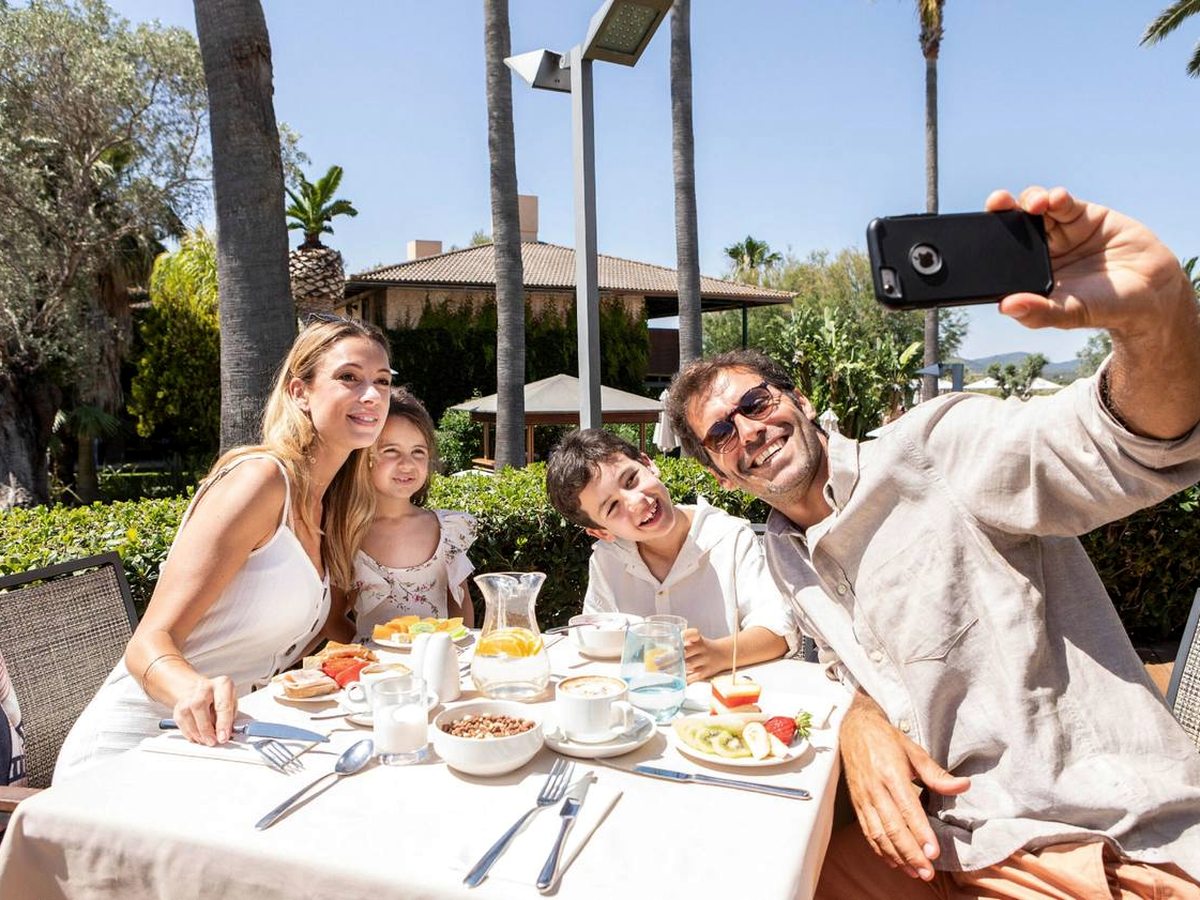 A family of four at an outdoor table laid with a white cloth, coffee, juice, fruit and cereal, with palms, hedges and a tiled resort building behind.
