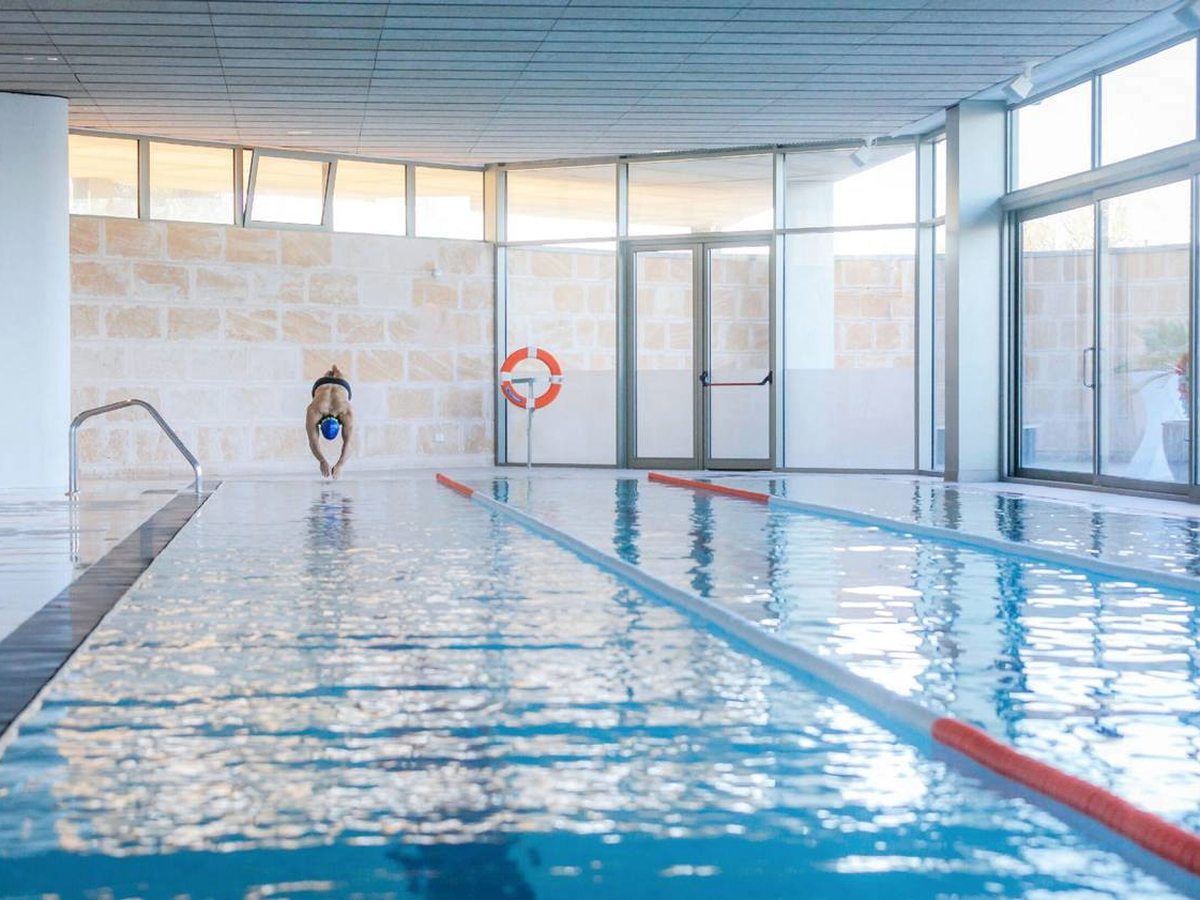 A long indoor lane pool with rope lane markers, a stone wall on one side and full-height windows on the other, with one swimmer in the water.