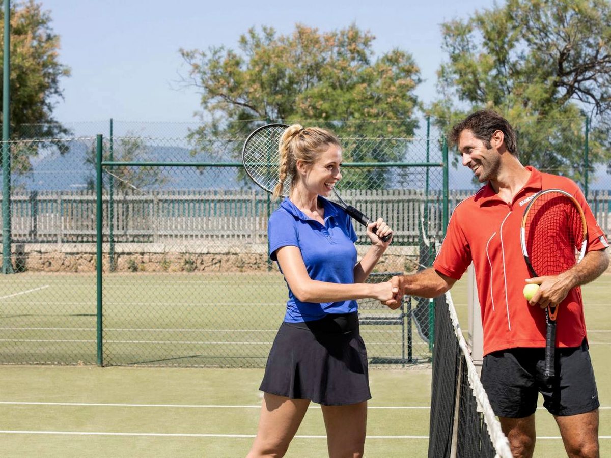 Two players shaking hands over the net on an artificial-grass tennis court, with a green fence, trees and hazy mountains behind.