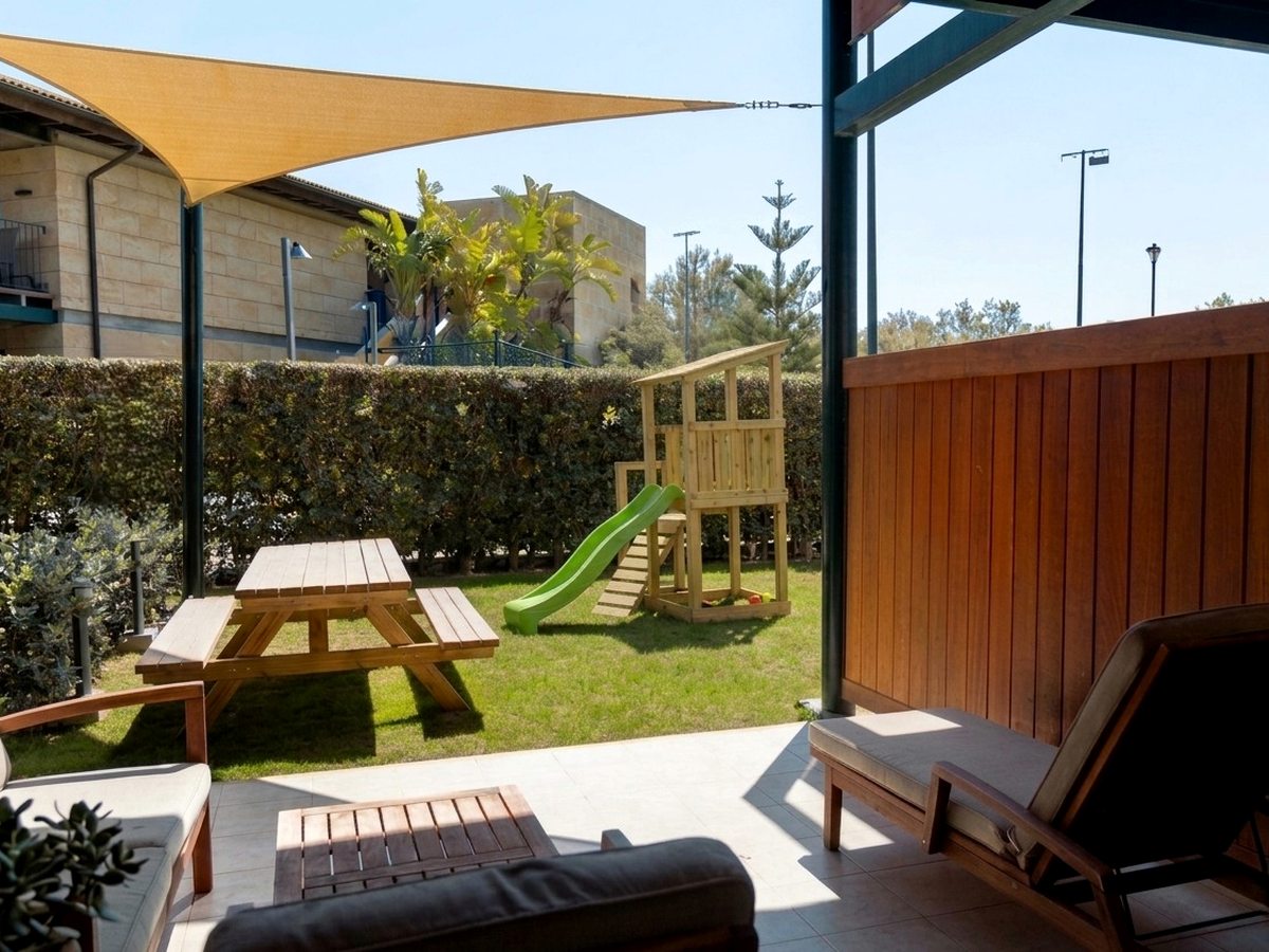 A hedged private lawn with a wooden climbing frame and green slide and a wooden picnic table, seen from the room's shaded terrace.