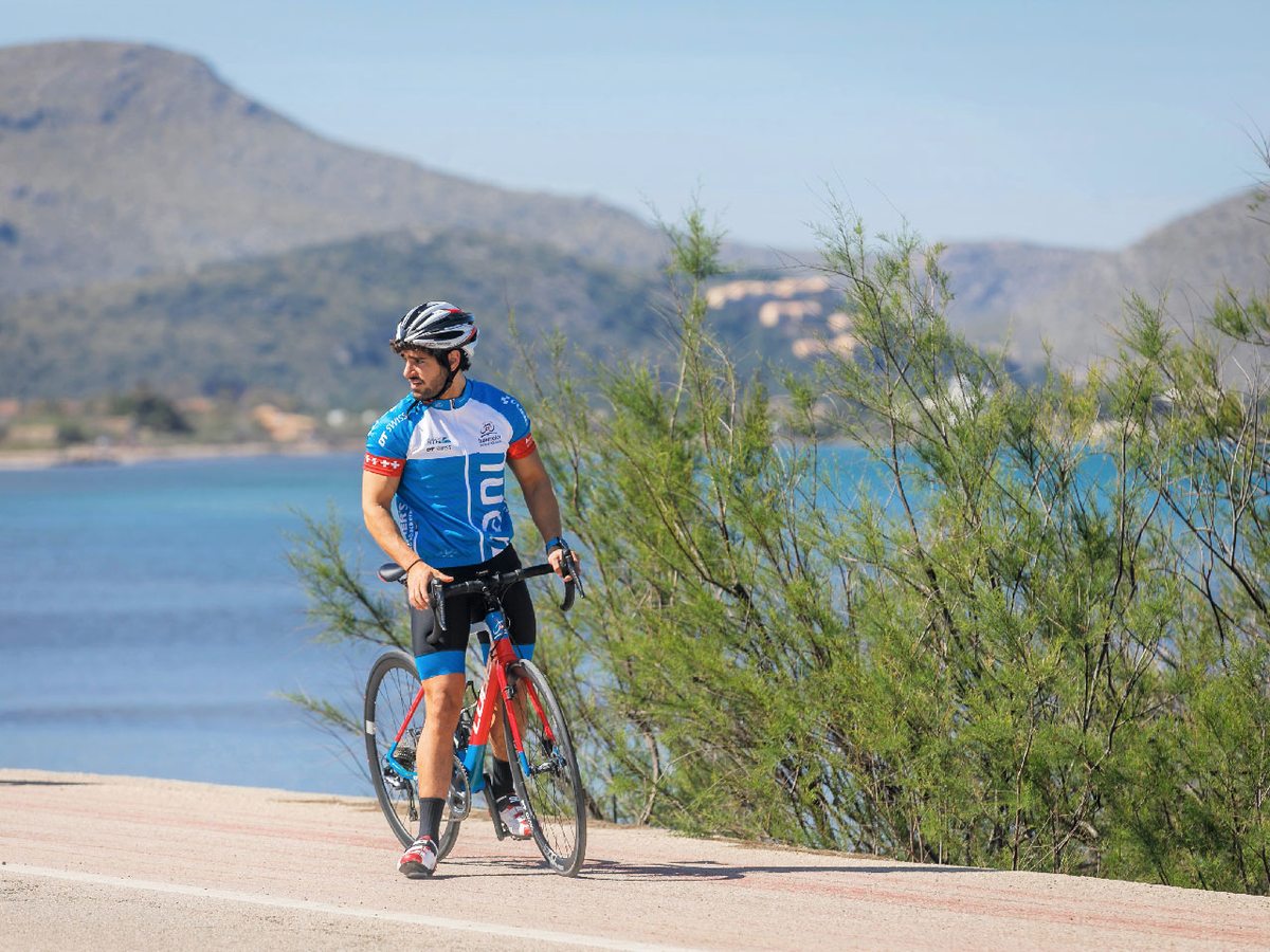 A road cyclist in team kit riding a coastal path beside the bay, with scrub in the foreground and a mountain headland across the water.