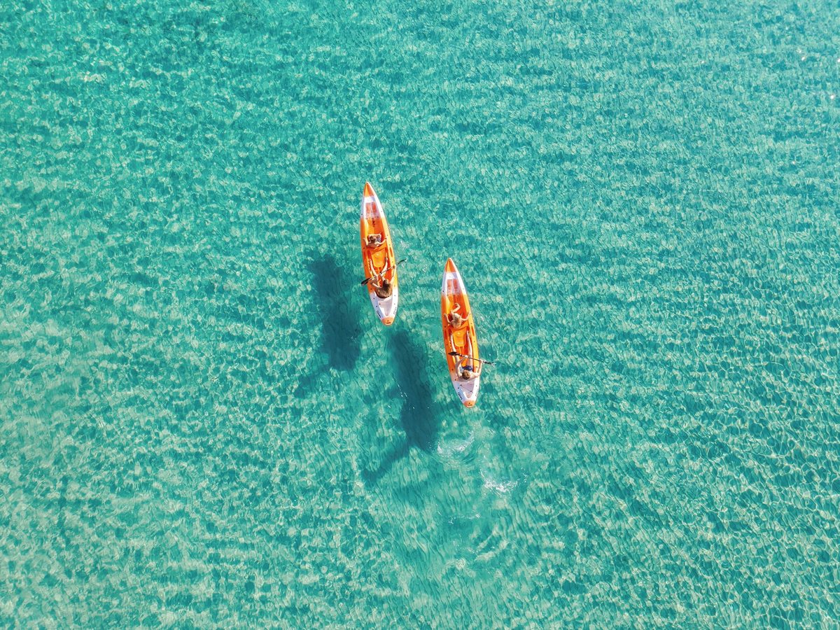 Two orange single kayaks being paddled across clear turquoise water with the pale seabed visible beneath.