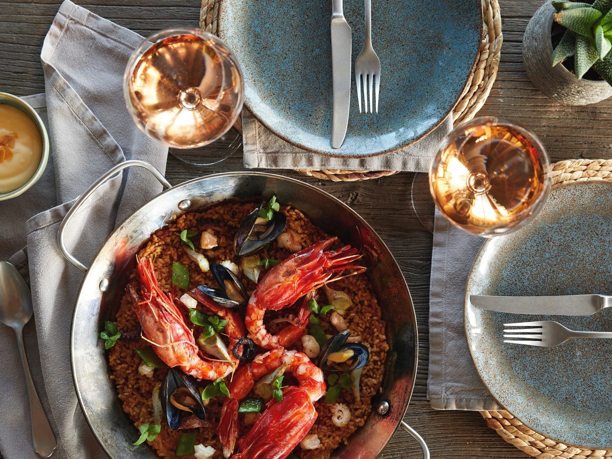 Overhead view of a seafood paella with whole prawns and mussels in a shallow pan, on a table with blue ceramic plates and cutlery.