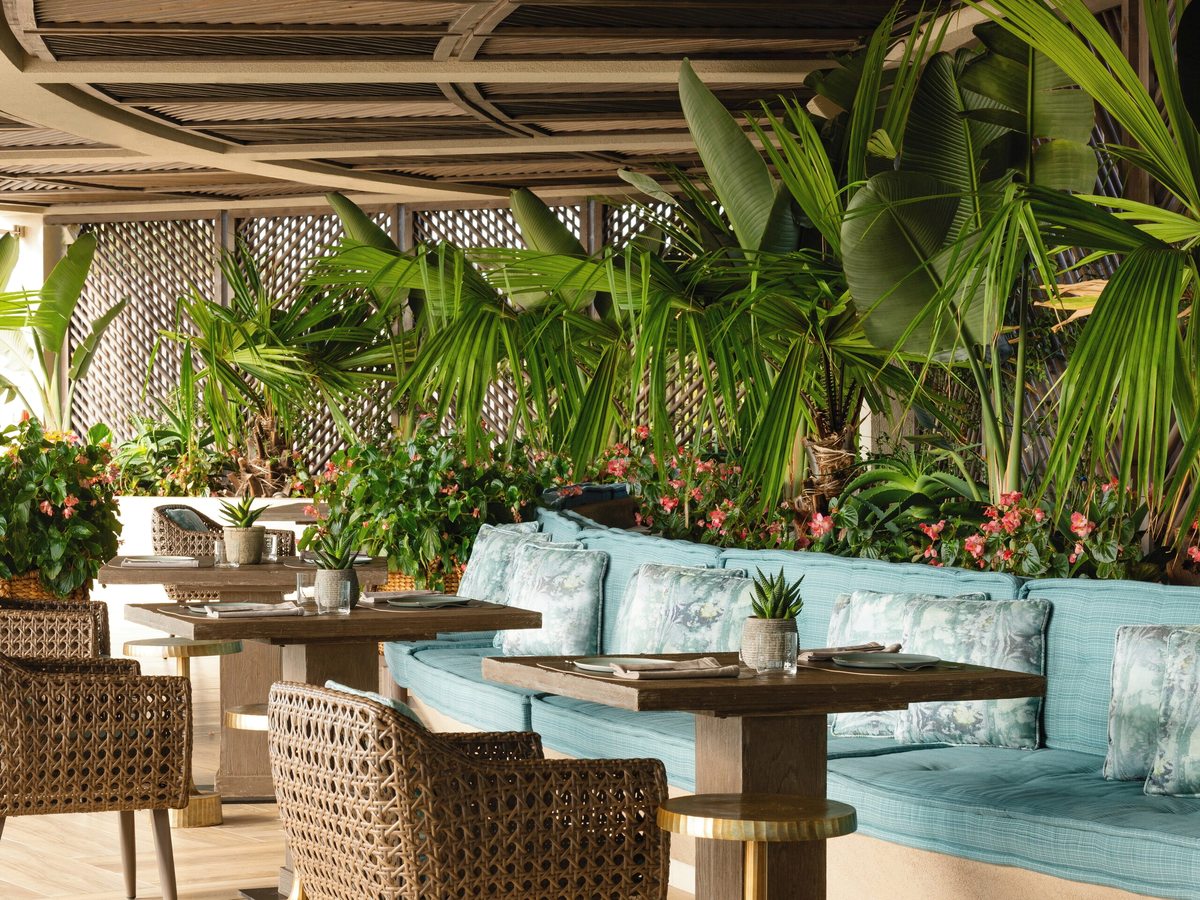 Covered restaurant terrace with wooden tables, woven armchairs and a long turquoise banquette, backed by dense palms and flowering plants under a timber pergola.