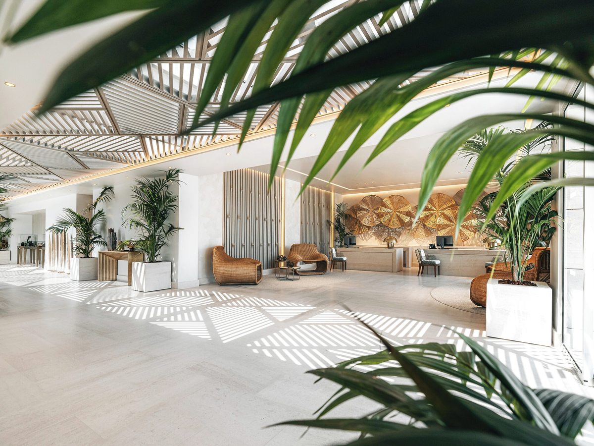 Bright open-plan hotel lobby with a pale stone floor, a slatted timber ceiling, tall potted palms and wicker armchairs in front of a gold fan-patterned wall behind the reception desk.