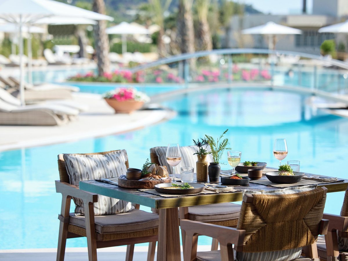 Outdoor table laid with plates, bread and wine glasses on the terrace at the edge of a turquoise lagoon pool, with sun loungers and parasols on the far side.