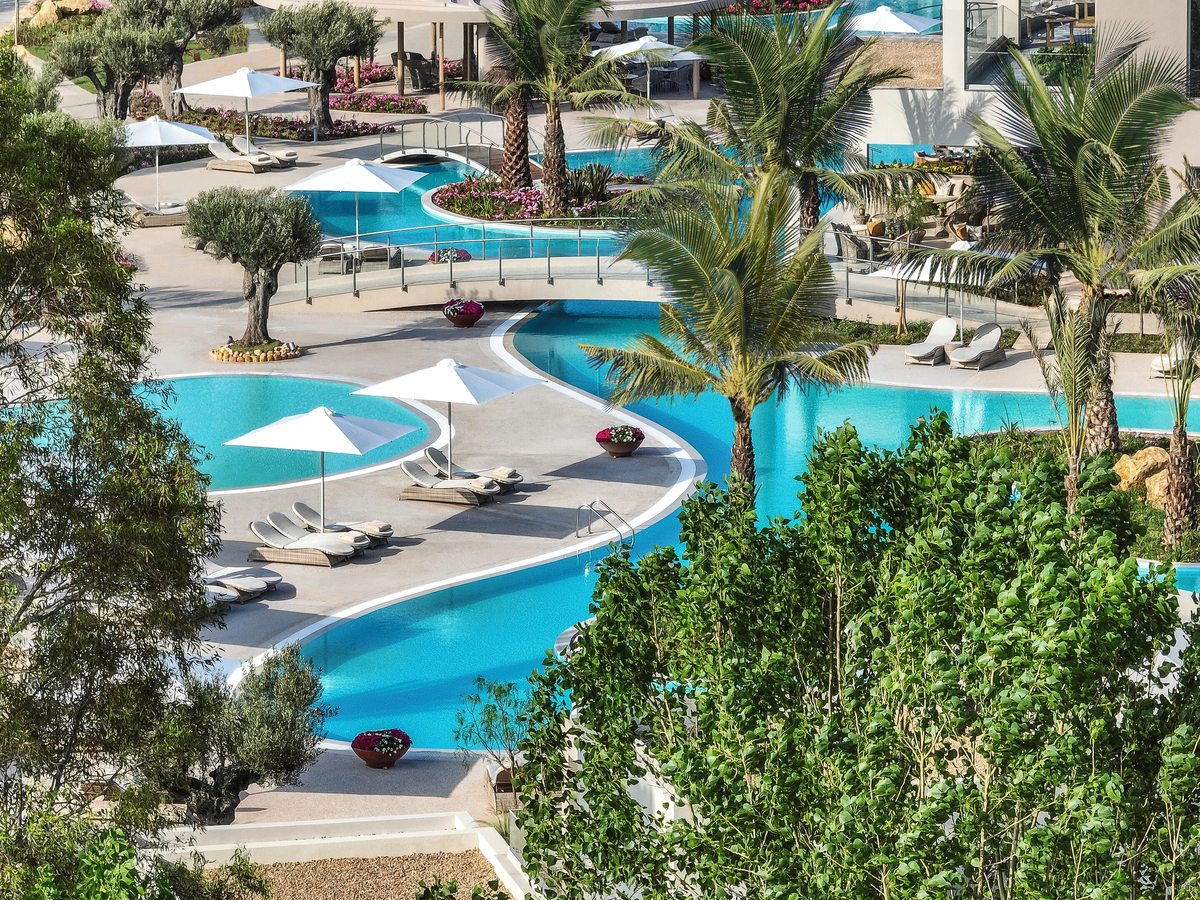 Elevated view of interlinked lagoon pools winding between planted terraces, with sun loungers, white parasols and olive trees around them.
