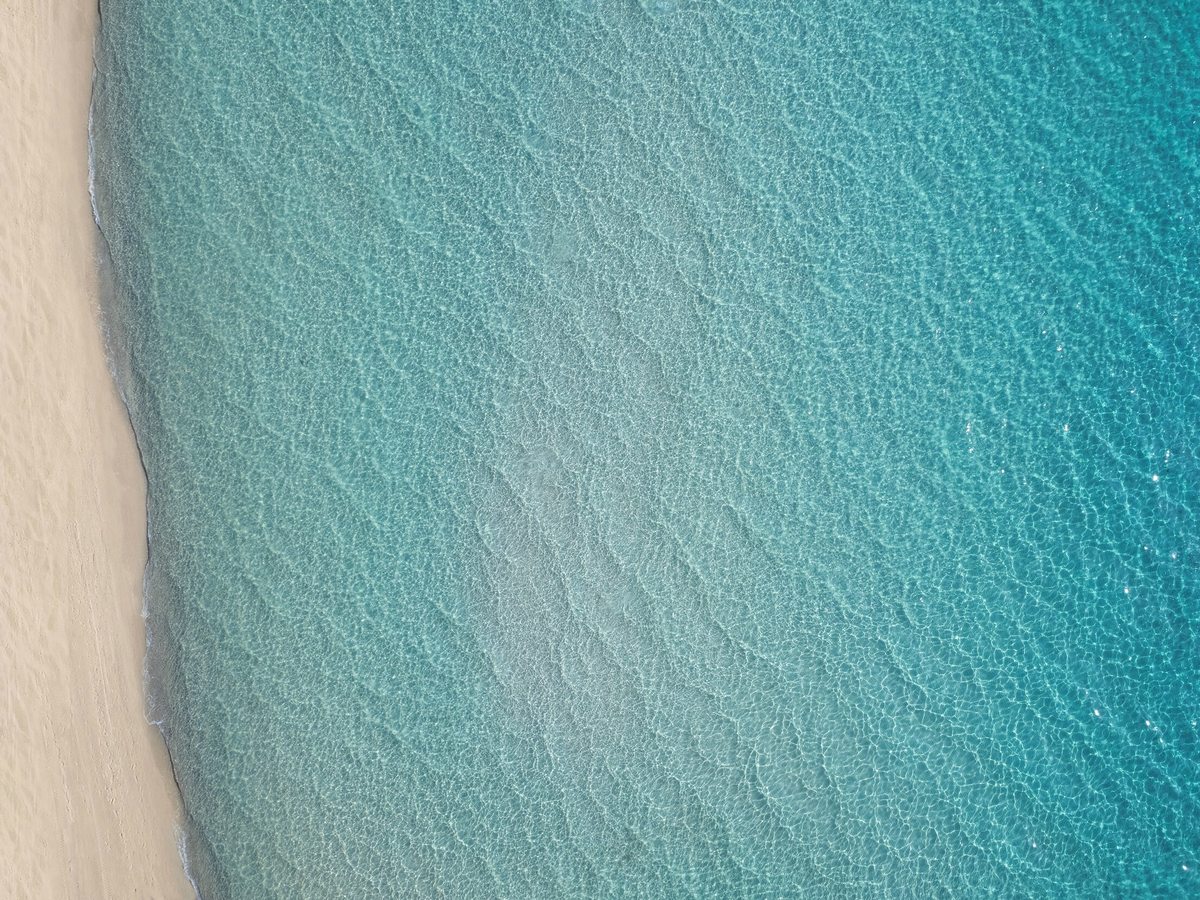 Overhead view of pale sand meeting clear turquoise water, shallow and rippled at the shoreline and deepening to blue.