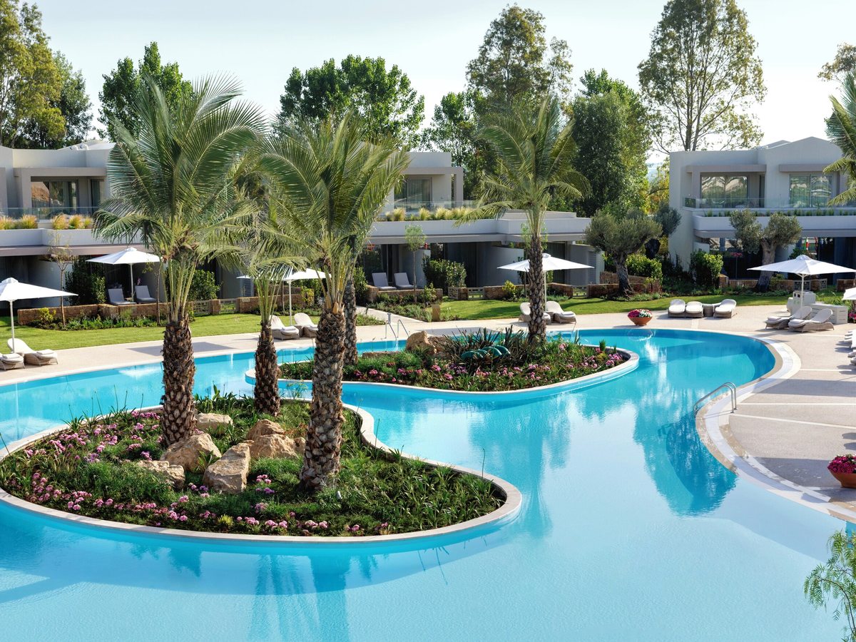 Curving turquoise lagoon pool with planted islands of palms and flowers, sun loungers and white parasols on the terrace and low white suite buildings behind.