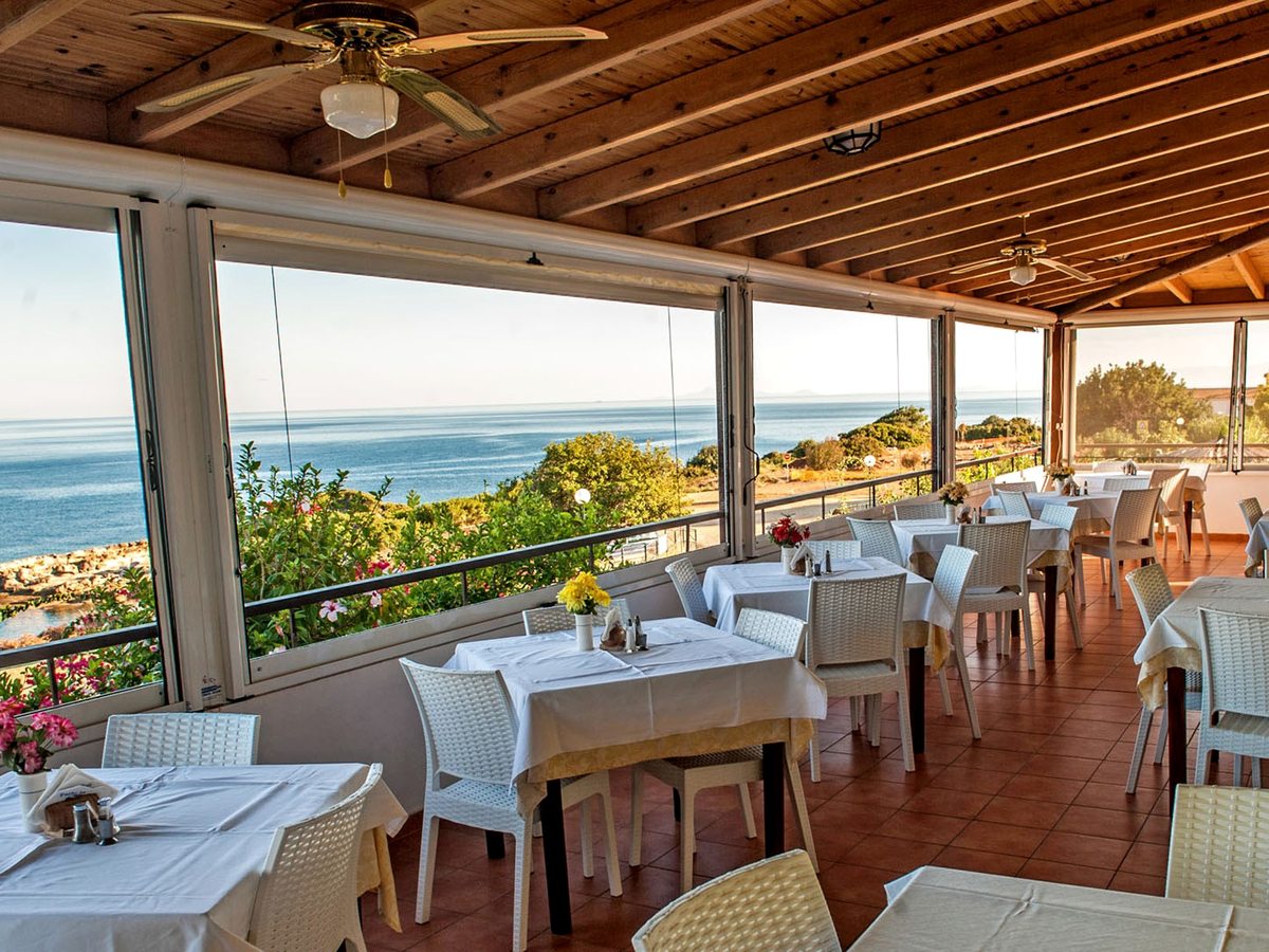 Covered restaurant terrace with a wooden ceiling and ceiling fans, white-clothed tables and pale wicker chairs, open along one side to the sea.