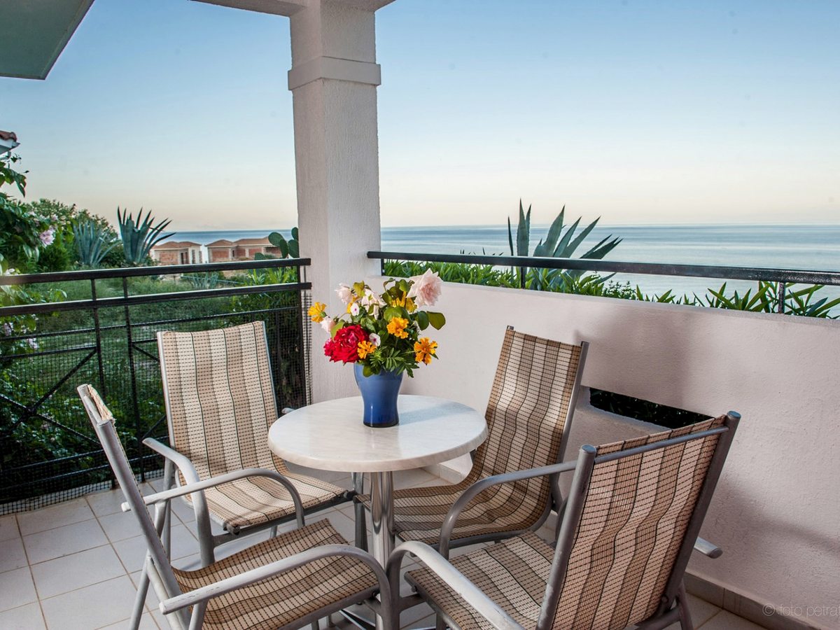 Covered balcony with a round table, four chairs and a vase of flowers behind a metal railing, with planting and the sea beyond.