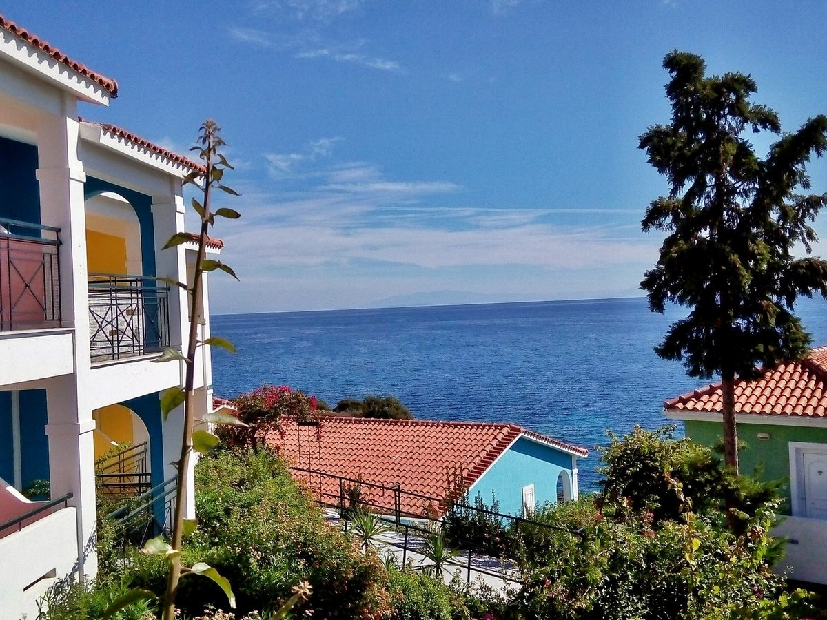 Two-storey pastel-painted accommodation blocks with balconies and shuttered doors, set among trees and shrubs under a clear blue sky.