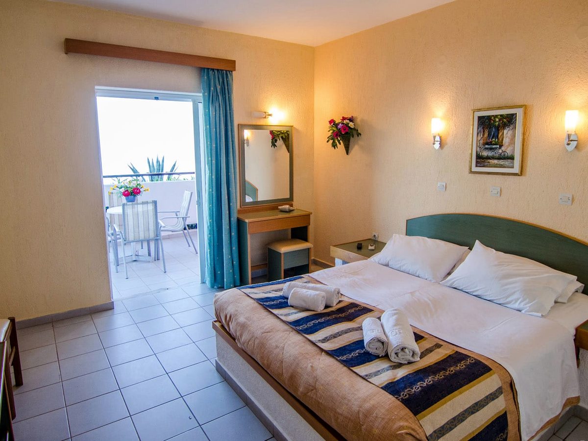 Hotel bedroom with a double bed, a dressing table and mirror, wall lights and open doors onto a balcony with a table and chairs.