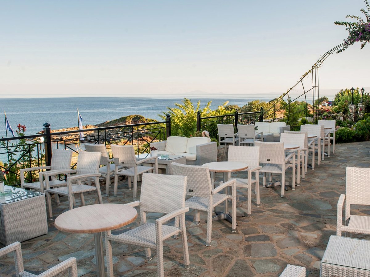 Stone-paved terrace set with white rattan tables, chairs and a cushioned sofa behind a black railing, with a rocky shore and calm sea beyond.