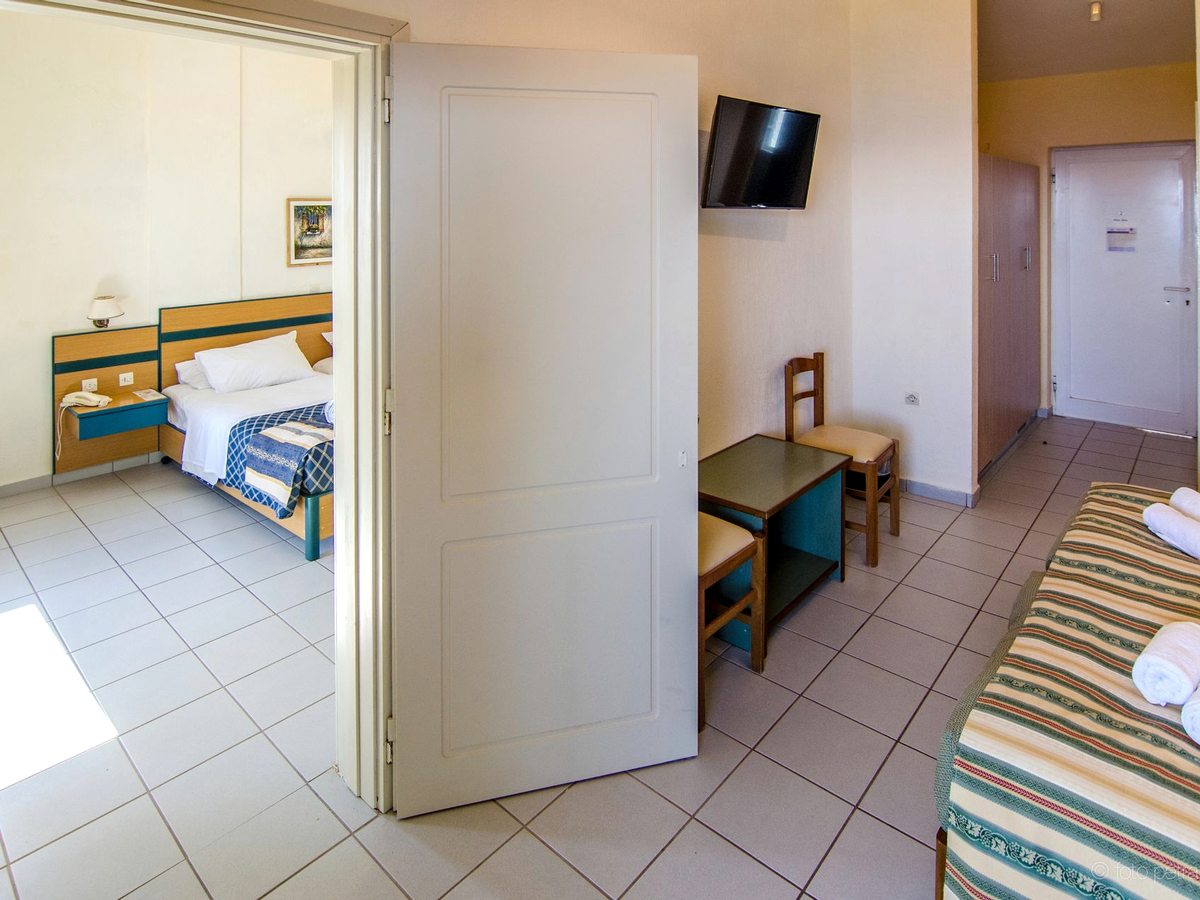 Hotel room with a bed, a table and chairs and a wall-mounted television, with a wide open door through to a second bedroom with a double bed.