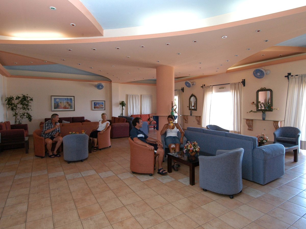 Indoor lounge with grey sofas and armchairs around low coffee tables on a tiled floor, with wall mirrors, potted plants and large windows.