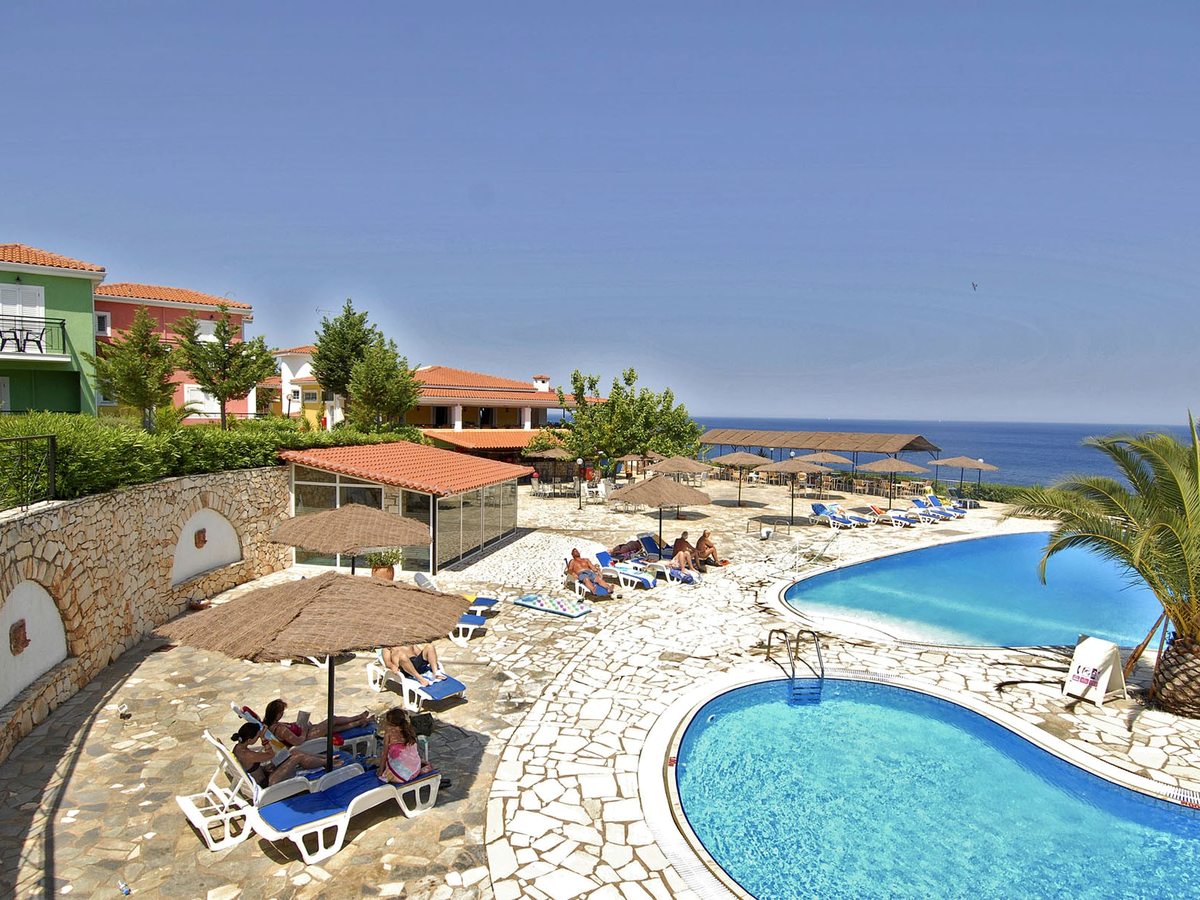 Guests on sun loungers under thatched parasols on a stone pool terrace, with two linked pools, a tiled-roof building and the sea beyond.