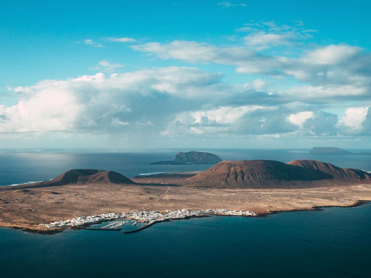 Aerial view of a low volcanic island with several brown cones and a white village on its harbour, with more islands out in the blue Atlantic beyond.