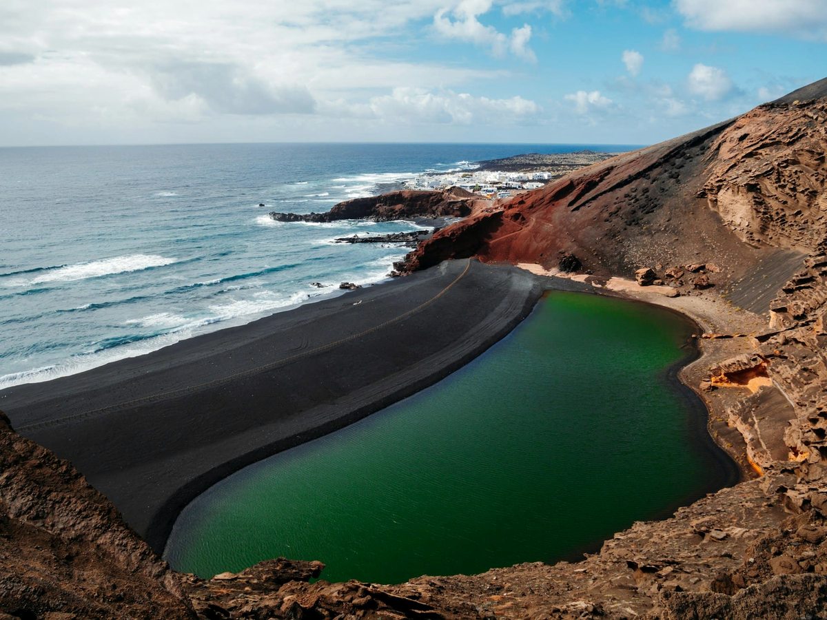 A bright green lagoon separated from the Atlantic by a curve of black sand, backed by red and ochre volcanic cliffs.