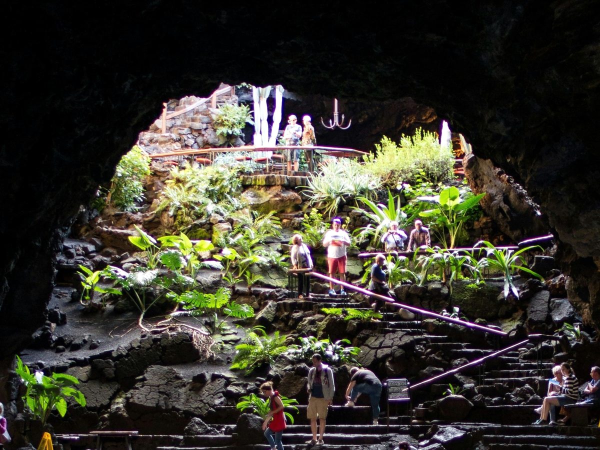 Visitors on stone steps and walkways inside a lava tunnel planted with palms and tropical foliage, lit by daylight through the open roof.