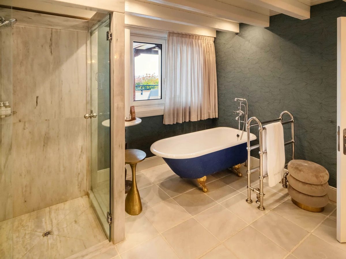 A bathroom with a blue roll-top bath on brass feet beneath a curtained window, a glass-walled walk-in shower and a dark slate wall.